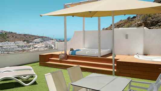 Terrass med solstolar, bord, parasoll samt utsikt över ett kuperat område med byggnader. (AI-genererad text)