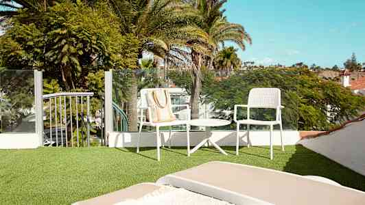 En terrass med grön syntetisk gräsmatta, möbler med vita stolar och ett bord samt utsikt över frodiga palmer mot en klar himmel. (AI-genererad text)