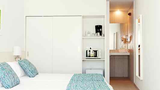 Ett hotellrum med en säng, lampor, och ett minikök som inkluderar en mikrovågsugn och en kaffemaskin. (AI-genererad text)