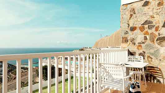 Utsikt från en balkong med ett bord och stolar, och en vy över havet och omgivningar. (AI-genererad text)