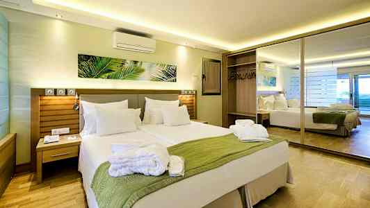 Ett hotellrum med en dubbelsäng, en grön filt och vita kuddar, samt en spegel och garderob i bakgrunden. (AI-genererad text)