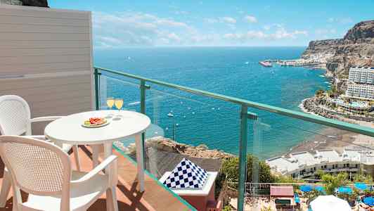 Utsikt från balkong med bord och två stolar, där glas serveras, mot havet och kustlandskap. (AI-genererad text)