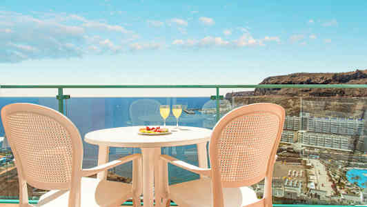 Bild av en balkong med utsikt över havet och omgivande byggnader. På balkongen står ett bord med två glas och en tallrik med frukt samt två stolar. (AI-genererad text)