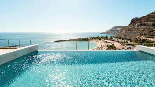 Utsikt över en infinitypool med solblänk på vattenytan och en sandstrand i bakgrunden. (AI-genererad text)