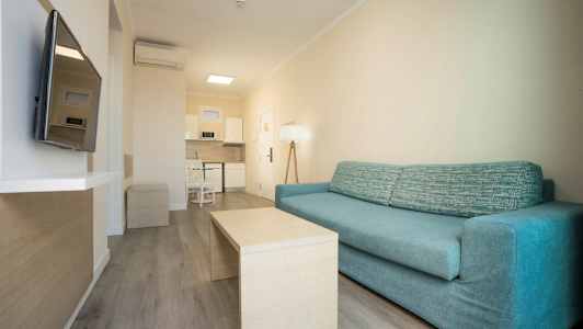 Ett vardagsrum med en blå soffa och ett bord framför, samt en TV på väggen och ett integrerat kök i bakgrunden. (AI-genererad text)