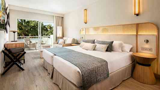 Ett hotellrum med en stor dubbelsäng, sittgrupp och balkong med utsikt mot gröna palmer. (AI-genererad text)