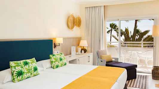 Ett hotellrum med en dubbelsäng som har gul sängöverkast och gröna prydnadskuddar, ljus inredning samt en balkong med utsikt mot palmer och havet. (AI-genererad text)