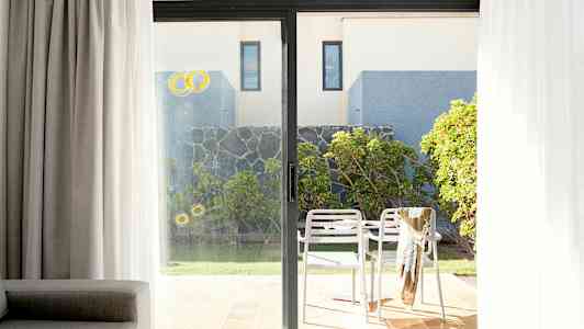 Bild av en uteplats med två vita stolar och ett bord, omgiven av grönska och en stenmur, sedd genom ett glasparti. (AI-genererad text)