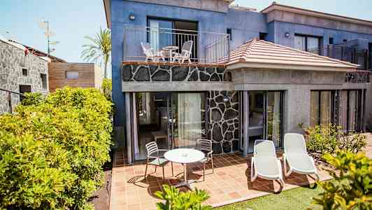 Bilden visar en hotellbyggnad med blå fasad och stenpanel. På markplan finns en terrass med utemöbler och solstolar, omgiven av grönska. (AI-genererad text)