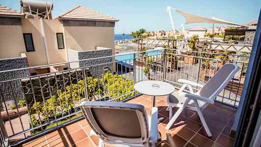 En balkong med två stolar och ett bord, utsikt mot byggnader och en pool. (AI-genererad text)