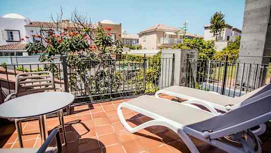 Bilden visar en balkong med ett bord, två stolar och två solstolar framför en räcke, med utsikt över en innergård och omgivande byggnader. (AI-genererad text)