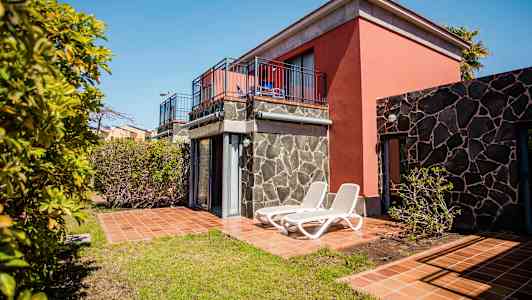 En semesterlägenhet med terrass och solstolar, omgiven av träd och grönska. (AI-genererad text)