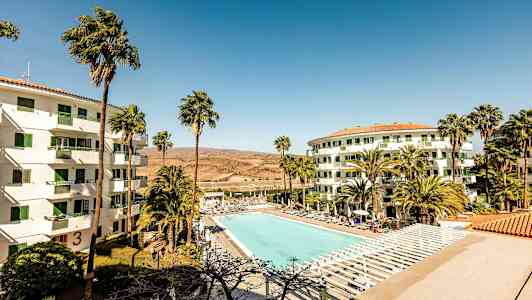 Bild av ett hotellområde med en stor utomhuspool omgiven av solstolar. Två vita byggnader med gröna fönster har palmer i förgrunden. (AI-genererad text)