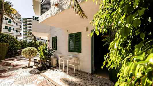 En uteplats med bord och stolar framför en vit byggnad med gröna fönsterluckor. Omgiven av träd och buskar. (AI-genererad text)