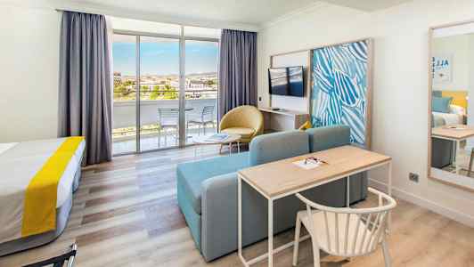 Ett hotellrum med säng, skrivbord, soffa, tv och glasdörrar som leder till en balkong. (AI-genererad text)