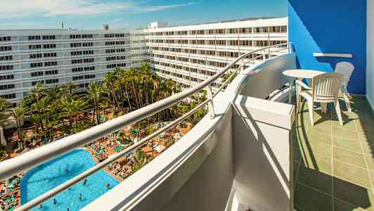 Utsikt från en balkong mot en poolområde med många solstolar och grönska, omgiven av vita hotellbyggnader. (AI-genererad text)