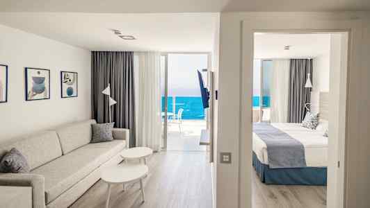 Vardagsrum med soffa och bord intill ett sovrum med dubbelsäng. Havsutsikt från balkongen. (AI-genererad text)