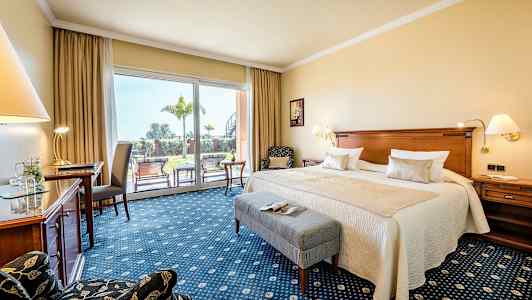 Ett hotellrum med en dubbelsäng, skrivbord, stol och stora glasdörrar som leder ut till en grönskande terrass. (AI-genererad text)