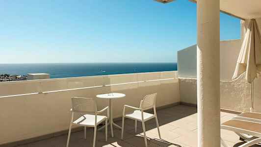 Uteplats med två vita stolar och ett bord, havet syns i bakgrunden. (AI-genererad text)