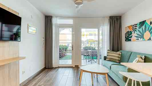 Ett ljust vardagsrum med en grön soffa, ett soffbord och en utsikt mot en terrass genom ett glasfönster. (AI-genererad text)