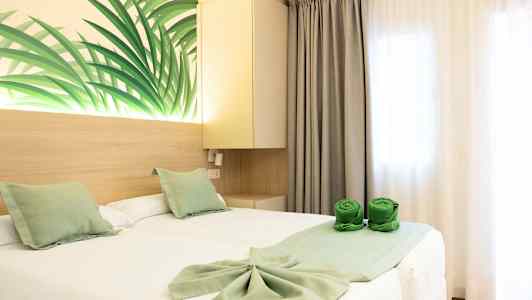 Ett hotellrum med två enkelsängar täckta av vita lakan och gröna dekorativa kuddar. Väggdekoration med bladmotiv samt gröna hoprullade handdukar på sängen. (AI-genererad text)