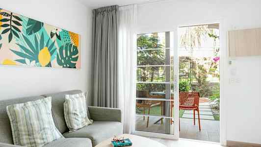 Vardagsrumsmiljö med grå soffa, prydnadskuddar, färgglad väggkonst, och glasdörr som leder till uteplats. (AI-genererad text)