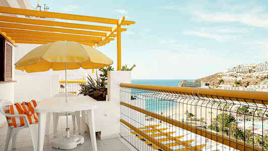 En balkong med utsikt över havet och stranden, möblerad med bord, stolar och ett gul parasoll. (AI-genererad text)