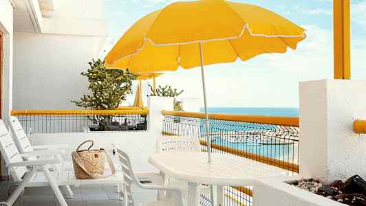 Vit uteplats med stolar, bord, gul parasoll och en havsutsikt. (AI-genererad text)