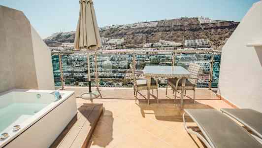 En terrass med utemöbler, ett parasoll och en bubbelpool med utsikt över en sluttning med byggnader. (AI-genererad text)