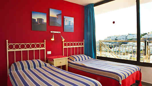 Två enkelsängar med randiga sängkläder och en röd vägg med doft bilder samt ett stort fönster med utsikt över byggnader. (AI-genererad text)