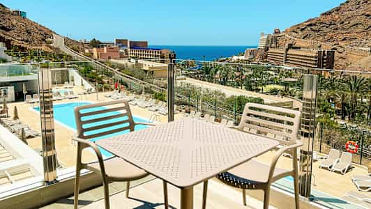 Utsikt över en pool och solstolar med havet och byggnader i bakgrunden, sett från en balkong med bord och stolar. (AI-genererad text)