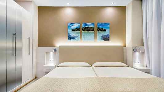 Bilden visar ett modernt hotellrum med en dubbelsäng, stilrena sidobord och en väggdekor med tre bilder av ett tropiskt strandmotiv. (AI-genererad text)