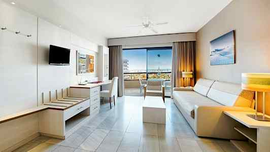 Ljusa hotellrum med soffor och möbler, öppet mot en balkong med havsutsikt. (AI-genererad text)