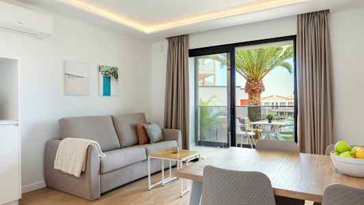 Ett vardagsrum med en grå soffa, ett soffbord och ett matbord. Utanför syns en terrass med utemöbler och en palm. (AI-genererad text)