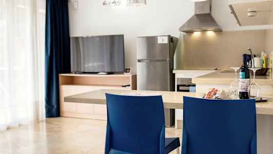 Ett kök med en diskbänk, två blå stolar, kylskåp, och en TV vid ett träskåp. (AI-genererad text)