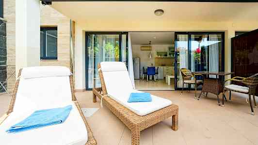 Två solstolar med vita dynor och en blå handduk på en terrass med bord och stolar framför glasdörrar till ett kök. (AI-genererad text)