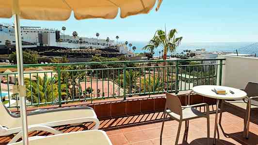 En terrass med bord, stolar och solstolar. I bakgrunden syns palmer, byggnader och havet. (AI-genererad text)