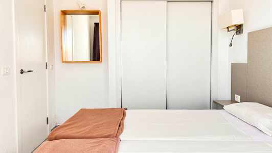 Ett hotellrum med vit inredning, en dubbel säng med vita lakan och bruna överkast, samt en spegel på väggen. (AI-genererad text)