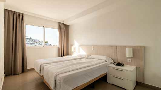 Ett hotellrum med en dubbelsäng och ett fönster som visar en vit byggnad. (AI-genererad text)