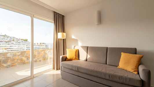 En grå soffa med gula kuddar står i ett rum med stora glasdörrar som leder ut till en balkong. (AI-genererad text)