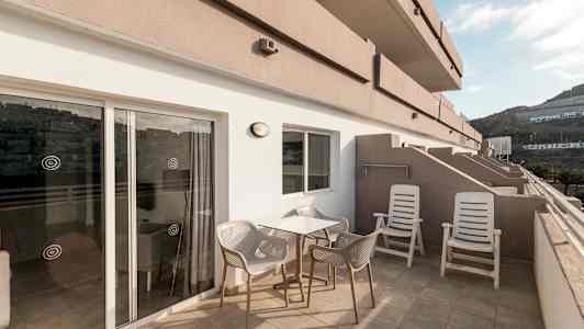 En terrass med bord, stolar och solstolar, samt utsikt mot omgivande byggnader och berg. (AI-genererad text)