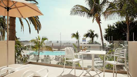 En balkong med solstolar och ett bord med stolar, utsikt över en pool och palmer. (AI-genererad text)