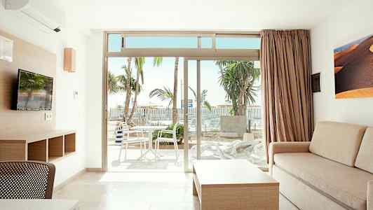 Ett ljust rum med soffa, bord och TV samt utsikt mot en terrass med bord och stolar. Palmträd syns utomhus. (AI-genererad text)