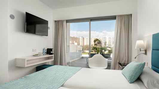 Ett hotellrum med två sängar bäddade med ljusa lakan och turkosa överkast. En stol står framför en glasdörr som leder ut till en balkong. En väggmonterad tv hänger bredvid en hylla med vattenflaskor och kaffemaskin. (AI-genererad text)