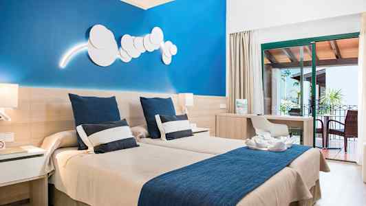 Ett hotellrum med två enkelsängar, en blå vägg, ett skrivbord och en möblerad balkong. (AI-genererad text)