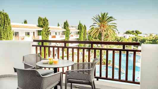 Utsikt från en balkong med ett bord och stolar där frukost är framställd. I bakgrunden finns en pool, grönområden och byggnader. (AI-genererad text)