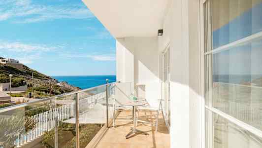 En balkong med glasräcke, ett bord och två stolar, med vy mot havet och omgivande landskap. (AI-genererad text)