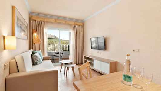 Ett vardagsrum med soffa, TV, bord och utsikt mot en balkong med stolar. (AI-genererad text)