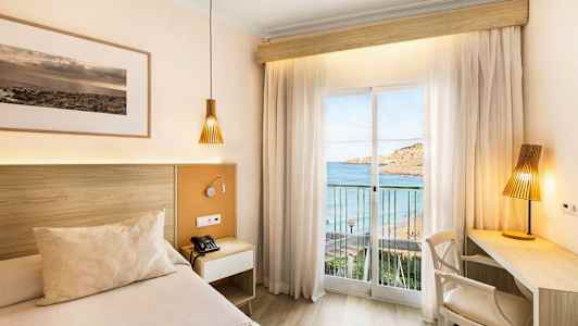 Ett hotellrum med en säng och skrivbord, med utsikt mot havet genom ett glasfönster med gardiner. (AI-genererad text)