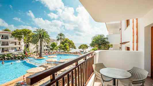 En utsikt över en balkong mot en pool omgiven av solstolar, palmträd och byggnader. (AI-genererad text)